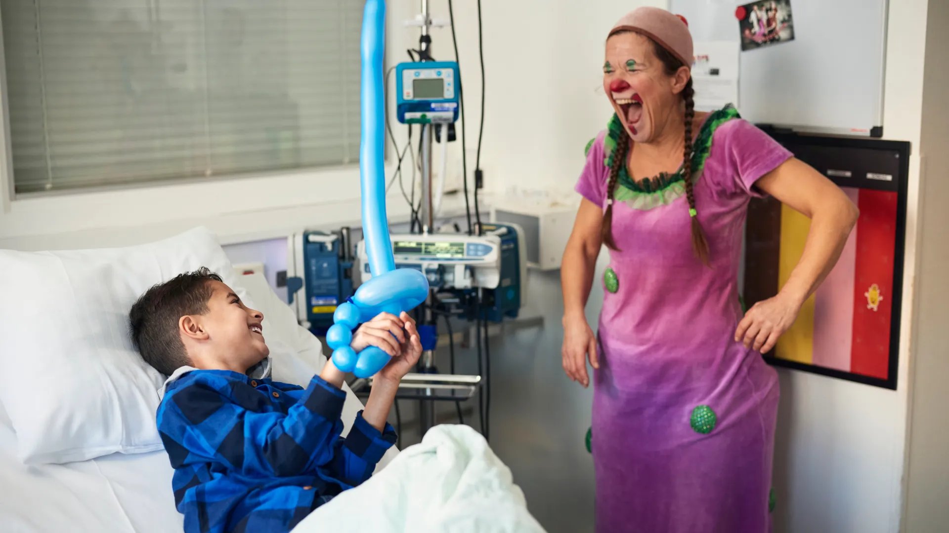Ett barn i en sjukhussäng leker med en clown i rosa klänning.